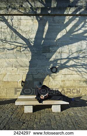 Stock Photo - A man lays across a bench reading a book; Paris, France. Fotosearch