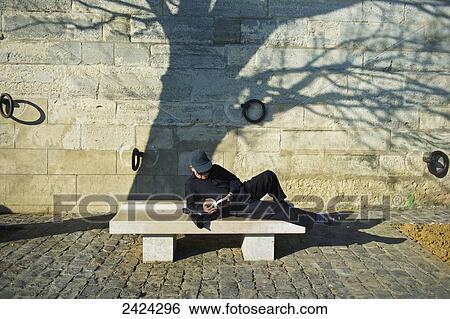 Stock Photograph - A man lays across a bench reading a book; Paris, France. Fotosearch