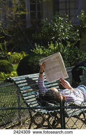 Stock Photo - A man lays on a bench reading a newspaper; Paris, France. Fotosearch