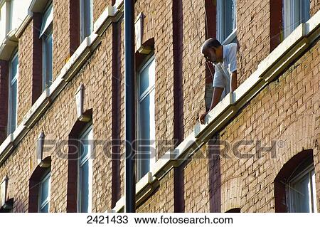 Stock Image - A man leans out the window of a brick residential building looking below; London, England. Fotosearch