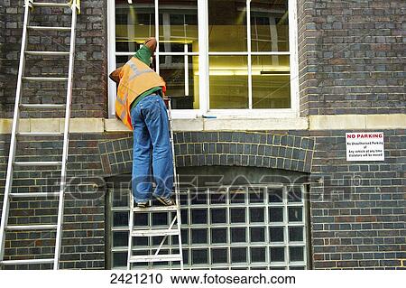 Stock Image - A man on a ladder repairing a window; London, England. Fotosearch