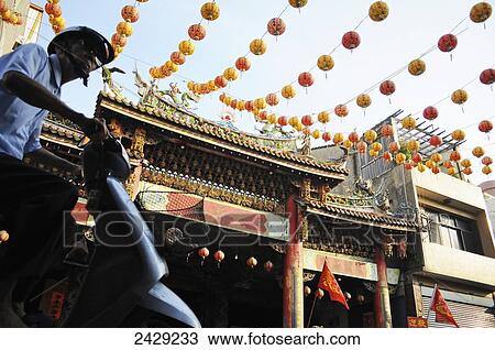 Stock Image - A man rides a scooter past a temple in downtown Lukang, a seaside city in Western Taiwan, and one of the few cities in the country that retain a traditional downtown core; Lukang, Taiwan. Fotosearch