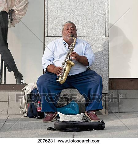 Stock Image - A Man Sits And Plays His Saxophone On The Sidewalk; Oslo Norway. Fotosearch