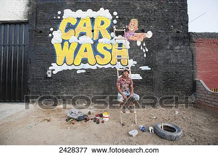 Stock Photo - A man sits on a ladder in front of his painted car wash sign on a brick wall; Chimbote, Peru. Fotosearch