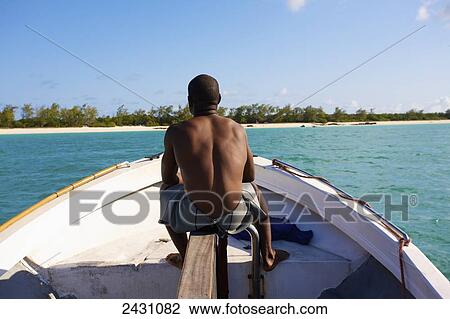 Stock Image - A man sits on his boat on the tranquil turquoise Indian Ocean with a view of the white sand beach; Vamizi Island, Mozambique. Fotosearch