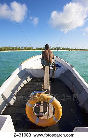 Stock Image - A man sits on his boat on the tranquil turquoise Indian Ocean with a view of the white sand beach; Vamizi Island, Mozambique. Fotosearch