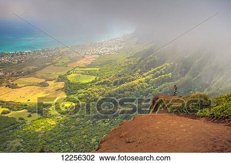 Stock Image - A man standing on the edge of a cliff on the Kuliouou Ridge Trial enjoys the view of Oahu's windward side and the town of Waimanalo as the clouds roll in on a summer's day; Waimanalo, Oahu, Hawaii, United States of America. Fotosearch