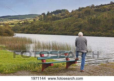 Picture - A Man Stands At A Bench Along The Shore Of Drinagh Lake; County Cork Ireland. Fotosearch
