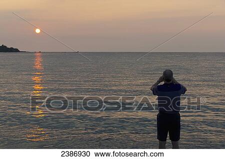 A man stands at the water's edge looking through binoculars at sunset in Anaehoomalu Bay; Island of Hawaii, Hawaii, United States of America View Large Photo Image Stock Image - A man stands at the water's edge looking through binoculars at sunset in Anaehoomalu Bay; Island of Hawaii, Hawaii, United States of America. Fotosearch