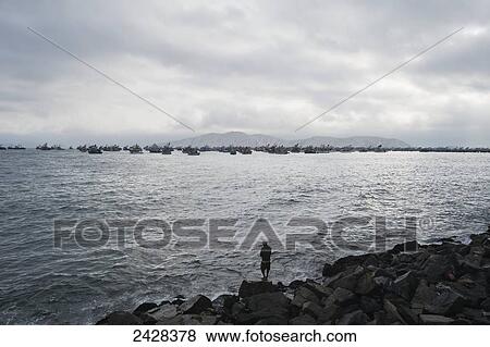 Stock Photo - A man stands on a rocky shore looking out at boats in a busy harbour; Chimbote, Peru. Fotosearch