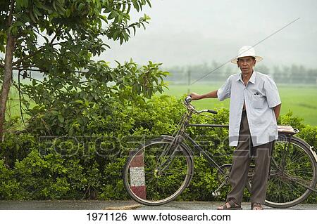Stock Photograph - A Man Stands With His Bicycle Beside A Lush Green Field; Mangshi Yunnan China. Fotosearch