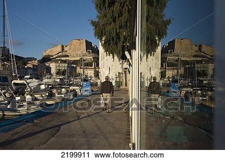 Stock Image - A man walks his dog in the early light of dawn. The citadel rises above the harbour of Bonifacio. Corsica. France. Fotosearch