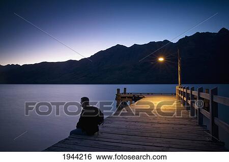 Stock Photograph - A Man Wearing A Backpack Sits On The Edge Of A Wooden Dock Illuminated With A Light At Dusk; Glenorchy South Island New Zealand. Fotosearch