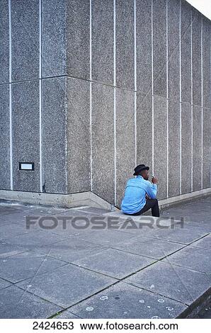 Stock Image - A man wearing a blue shirt and hat sits on a step beside a concrete wall, Southbank; London, England. Fotosearch