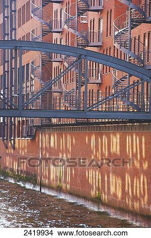 Picture - A metal footbridge crossing over a canal from a brick building with spiral staircases going up the exterior wall; Hamburg, Germany. Fotosearch