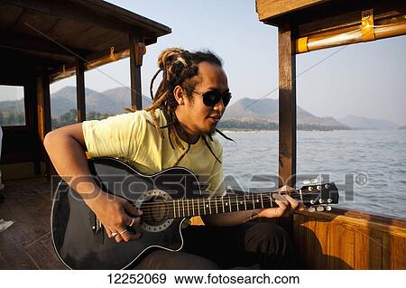 Stock Photo - A musician plays the guitar and sings on a river cruise along the Mekong outside of Luang Prabang; Laos. Fotosearch