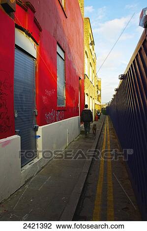 Stock Image - A pedestrian and dog walking down a narrow alley beside colourful buildings; London, England. Fotosearch