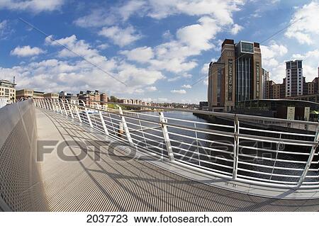 Stock Image - A Pedestrian Bridge Across River Tyne With Buildings Along The Waterfront; Newcastle Northumberland England. Fotosearch