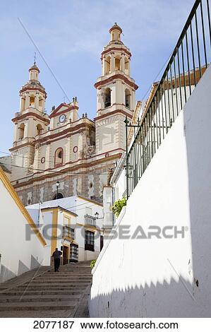 Stock Photo - A Pedestrian Walking Up The Steps Towards A Building With Two Towers And A Clock; Olvera Andalusia Spain. Fotosearch