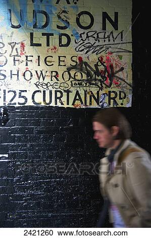 Stock Image - A pedestrian walks by a wall with graffiti over a sign painted on a wall, Shoreditch; London, England. Fotosearch