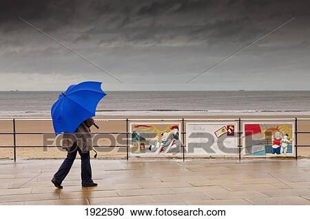 Stock Image - A Person Walks With An Umbrella On A Promenade Along The Water On A Rainy Day; Redcar, Teesside, England. Fotosearch