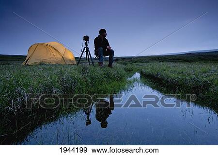 Stock Photo - A Photographer Sits By His Tent And A Stream Of Water; Iceland. Fotosearch