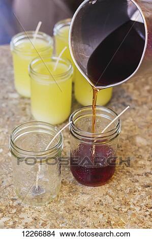 Picture - A pitcher is used to pour hot soy wax into glass mason jars with candle wicks for the final step in candle making for home made gifts; Fort McMurray, Alberta, Canada. Fotosearch