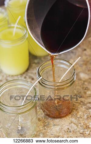 Stock Image - A pitcher is used to pour hot soy wax into glass mason jars with candle wicks for the final step in candle making for home made gifts; Fort McMurray, Alberta, Canada. Fotosearch