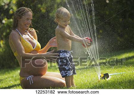 Stock Image - A pregnant mother in a bikini sits on the wet lawn and plays with her young son by the sprinkler; Santa Barbara, California, United States of America. Fotosearch