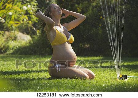 Stock Image - A pregnant woman in a bikini sits on the wet lawn by the sprinkler; Santa Barbara, California, United States of America. Fotosearch