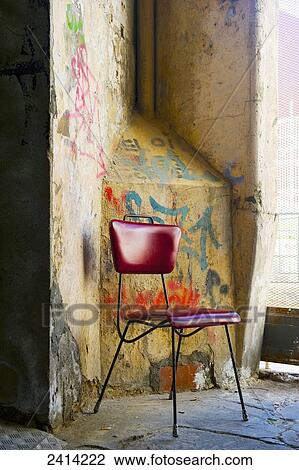 Stock Image - A red chair sits beside a weathered wall with painted graffiti; Barcelona, Spain. Fotosearch