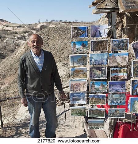 Stock Image - A Senior Man Stands Beside A Postcard Souvenir Rack; Nevsehir Turkey. Fotosearch