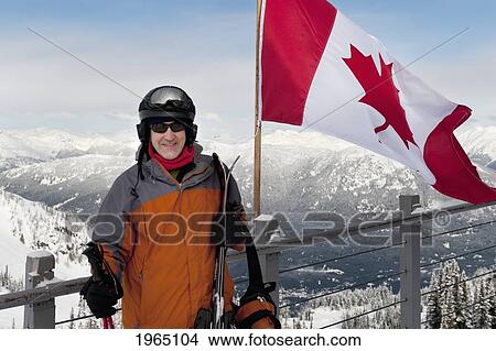 Picture - A Skier Stands Beside The Canadian Flag On A Balcony At A Ski Resort; Whistler British Columbia Canada. Fotosearch