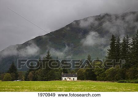 Stock Photo - A Small House Under Low Lying Clouds With Trees And A Mountain In The Background; Argyl Scotland. Fotosearch