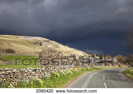 Stock Image - A Stone Wall And Buildings Along A Paved Road With Dark Storm Clouds Overhead; Muker Village Yorkshire England. Fotosearch