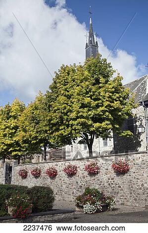 Stock Photograph - A Stone Wall Decorated With Hanging Flower Pots And The Spire Of A Church In The Background; Hargnies Ardennes France. Fotosearch