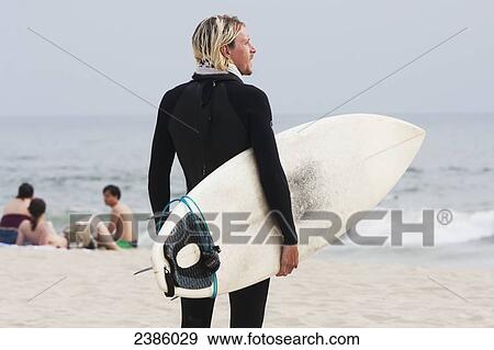 Stock Photo - A surfer stands on the beach holding his surfboard and looking out at the water; California, United States of America. Fotosearch