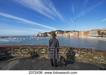 Stock Photograph - A Woman And Her Dog Stand At The Water's Edge Of The Harbour Looking Out Over The Coastline; Sestri Levante, Leguria, Italy. Fotosearch