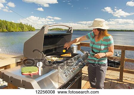 Stock Photo - A woman cooks a meal on a barbeque on a deck overlooking a beautiful lake; Ontario, Canada. Fotosearch