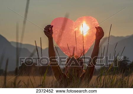 Stock Image - A Woman Holds Up A Red Heart With The Sun To Shine Through; Locarno, Ticino, Switzerland. Fotosearch
