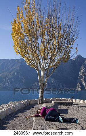 Stock Photo - A woman laying on a bench at the water's edge of Lake Garda; Malcesine, Verona, Italy. Fotosearch