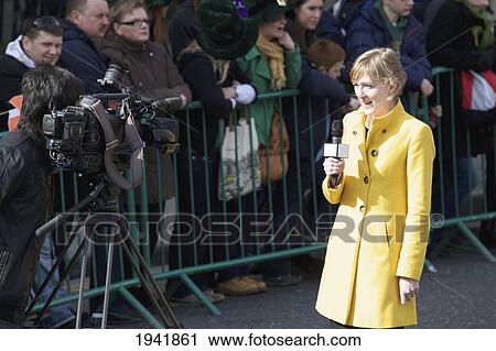 Stock Image - A Woman News Reporter Reporting In Front Of A Camera Beside The Crowd Waiting At The Saint Patrick's Day Parade; Dublin Ireland. Fotosearch