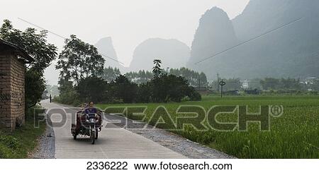 Stock Image - A Woman Rides A Motorbike With Cart Down A Path Beside A Field; Guilin, Guangxi, China. Fotosearch