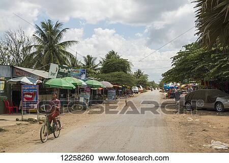 Stock Image - A woman rides her bicycle down a dirt road lined with shops and umbrellas; Battambang, Cambodia. Fotosearch