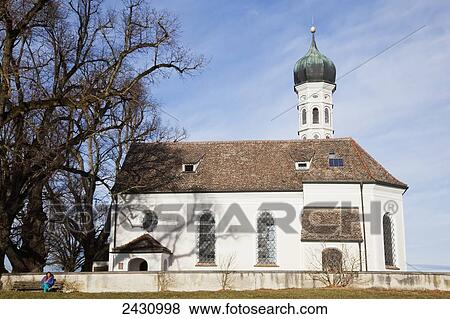 Stock Photo - A woman sits alone on a bench outside a church building; Bavaria, Germany. Fotosearch