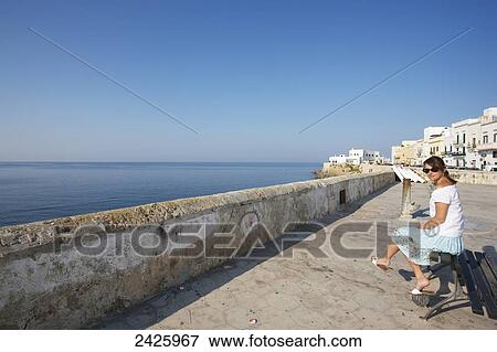 Stock Photo - A woman sits on a bench on a promenade overlooking the water, Gallipoli; Salento, Italy. Fotosearch