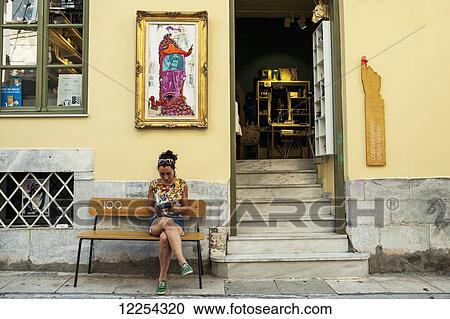 Stock Image - A woman sits on a bench outside a yellow building; Athens, Greece. Fotosearch