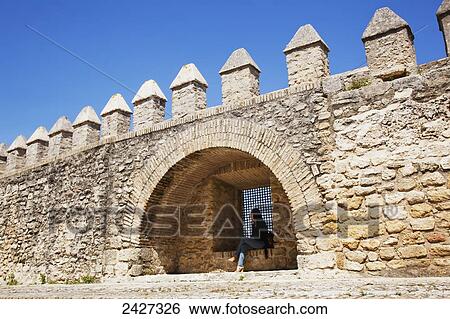 Stock Photograph - A woman sits on a ledge inside a niche of an old stone wall; Vejer de la Frontera, Andalusia, Spain. Fotosearch