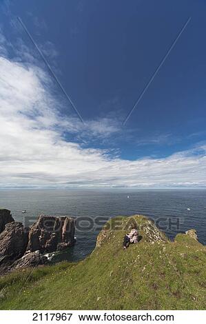Stock Photo - A Woman Sits On The Grass Along The Coast; St Abbs Head Scottish Borders Scotland. Fotosearch