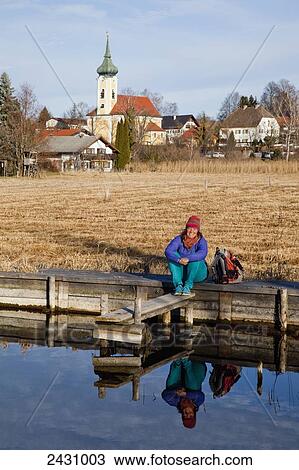 Stock Image - A woman sits smiling at the water's edge with the buildings of Murnau in the background; Seehausen, Bavaria, Germany. Fotosearch